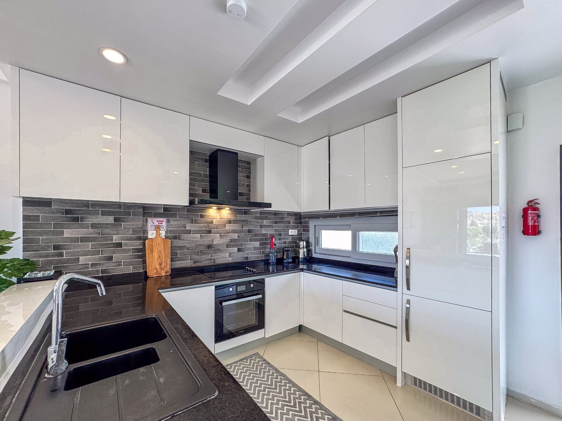 Modern L-shaped kitchen with white high-gloss cabinetry, dark stone counters and built-in oven