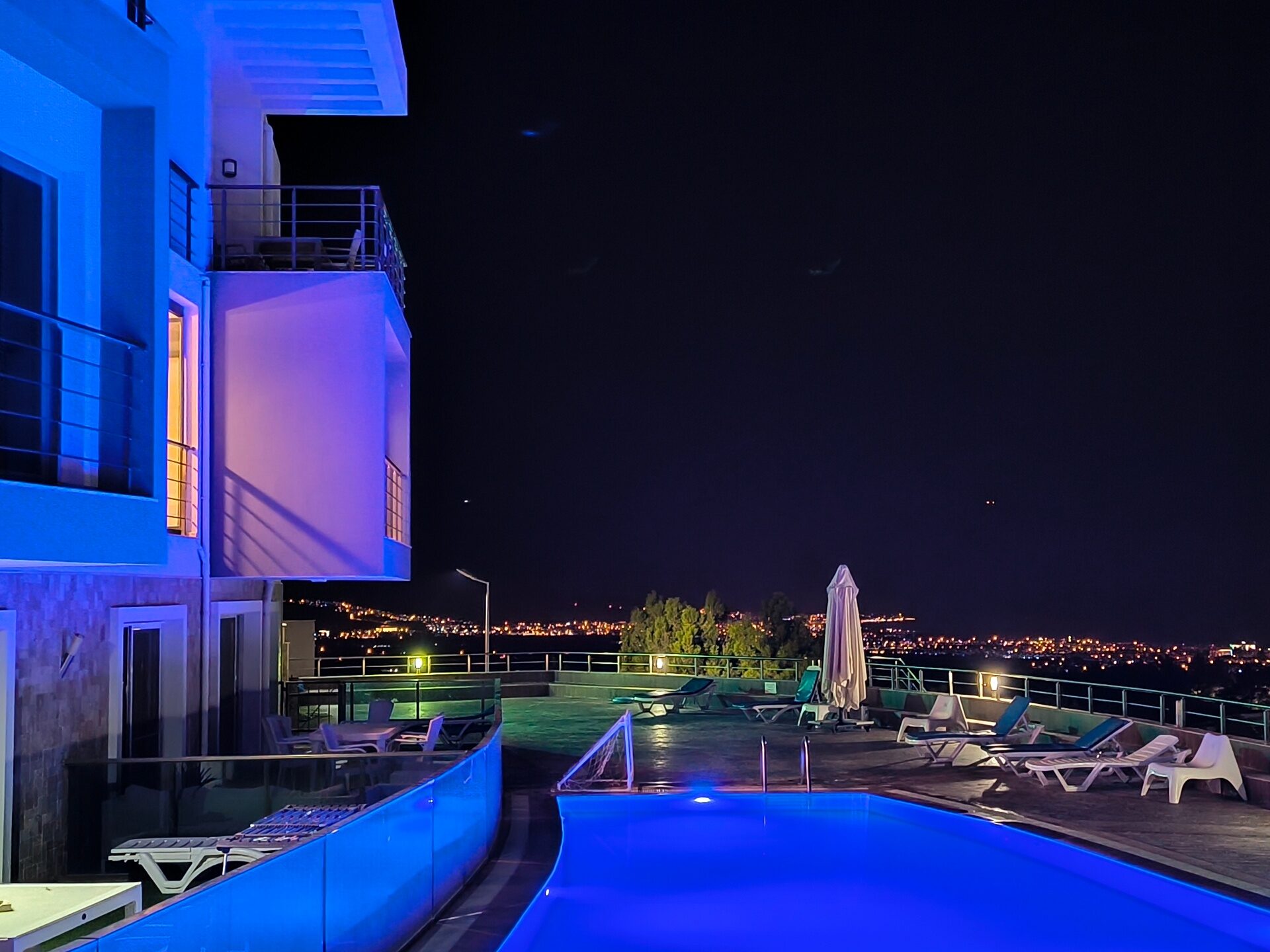Swimming pool and terrace lit blue at night with the city lights below